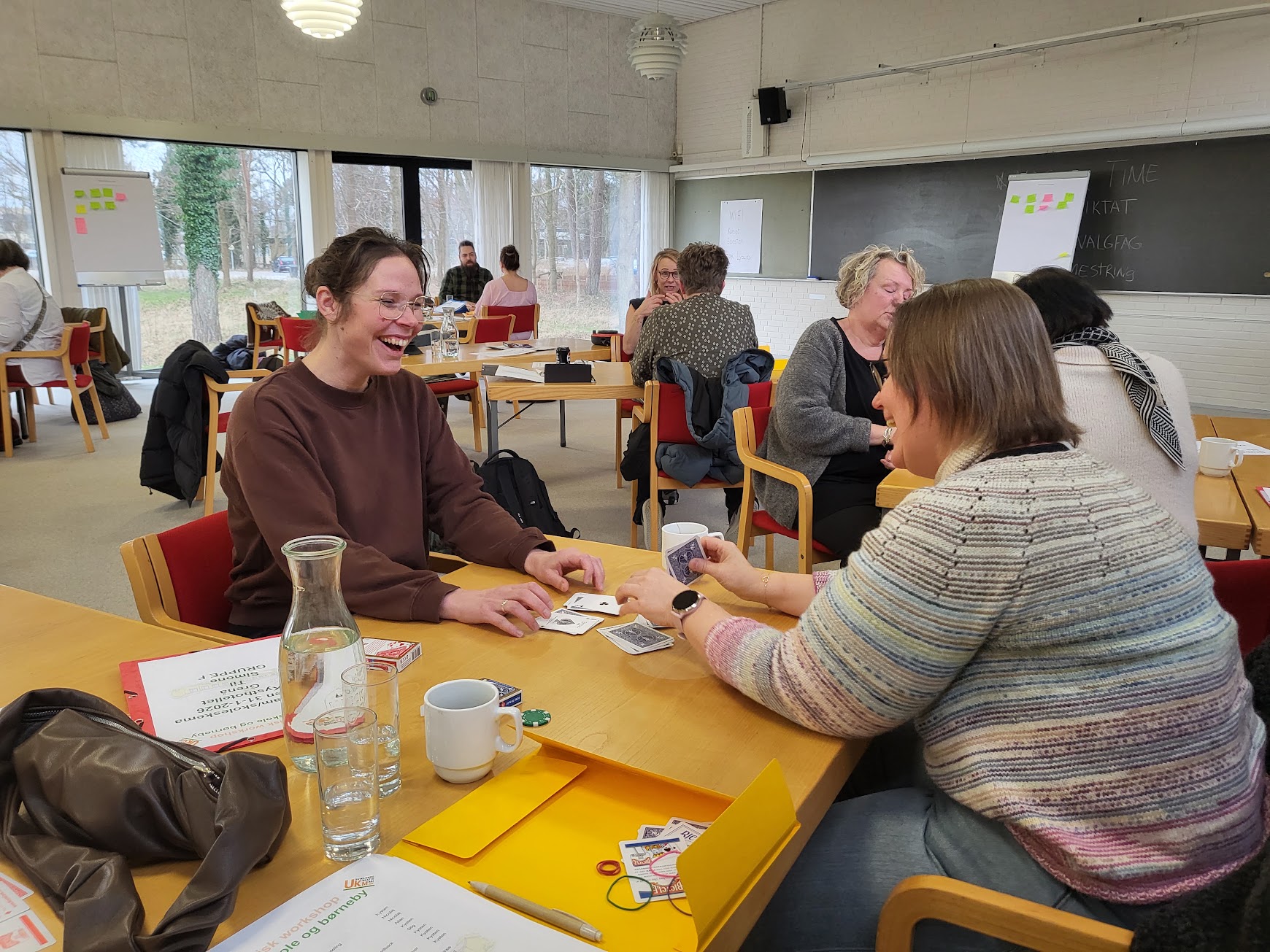 Deltager griner, mens en kollega viser et korttrick ved bordet under magisk teambuilding